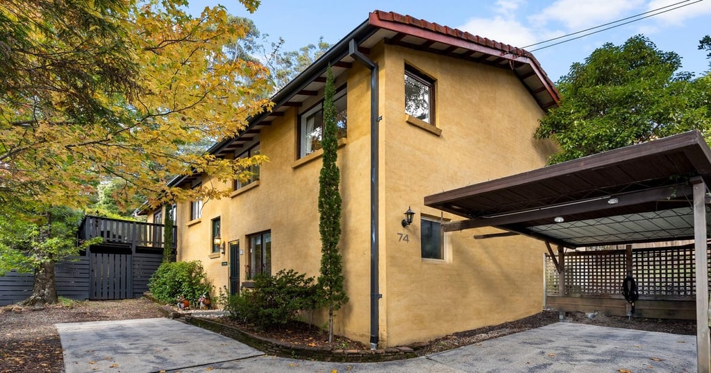 Ochre two-storey home at 74 Leura Mall, with a carport, driveway, garden trees and the number 74 beside the entrance